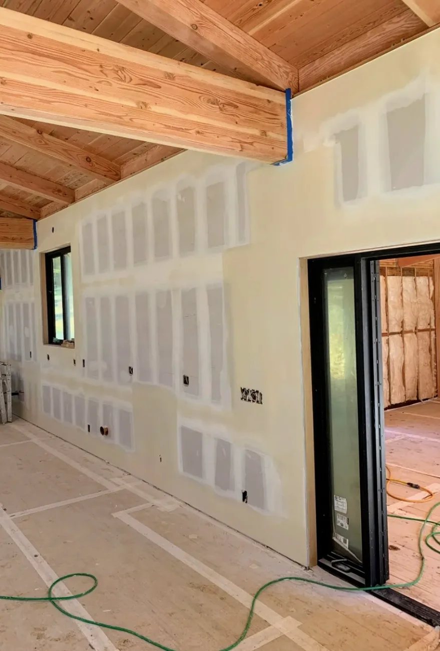 Newly hung drywall taped and coated over every seam and screw, fitted around exposed timber beams and a black-framed door