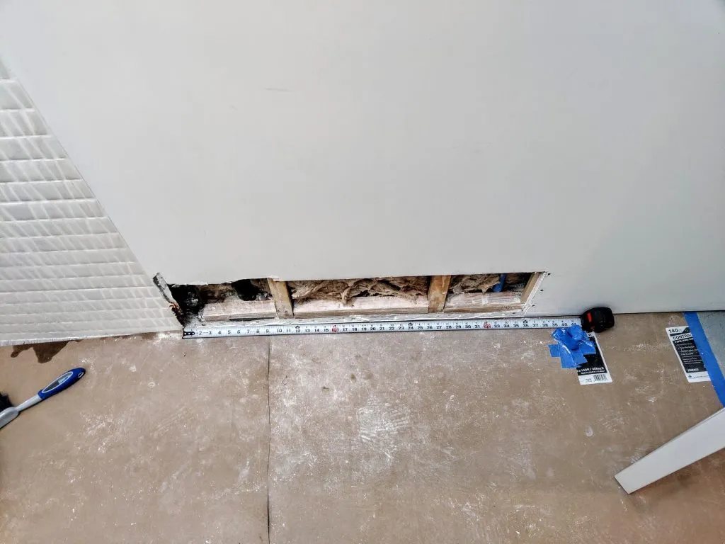 Water-damaged drywall cut back at floor level, exposing studs and wet insulation, measured out before new board goes in